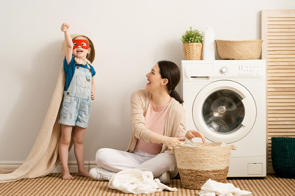 Mutter sitzt neben Waschmaschine und ein kleiner Junge steht wie Supermann verkleidet neben ihr