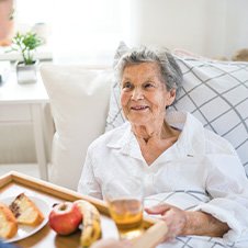 Pflegekraft bringt einer älteren Dame im Bett das Frühstück