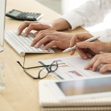 Bildausschnitt: Geschäftsleute an einem Tisch mit Notebook,Brille, Taschenrechner und Unterlagen