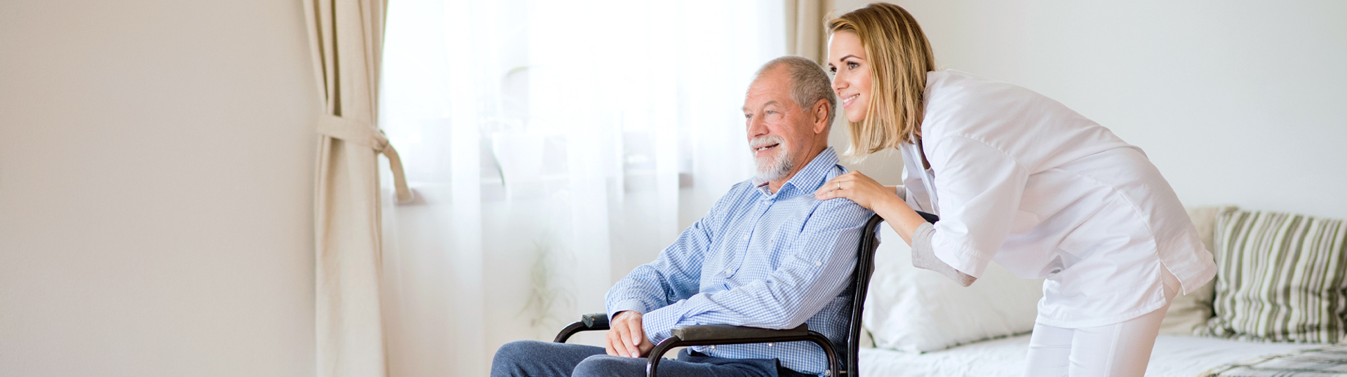 Pflegerin beugt sich zu älterem Mann im Rollstuhl und beide lächeln