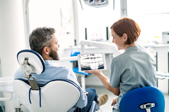 Zahnärztin sitzt neben einem Patienten im Behandlungsstuhl und erklärt ihm die Behandlung mit einem Tablet