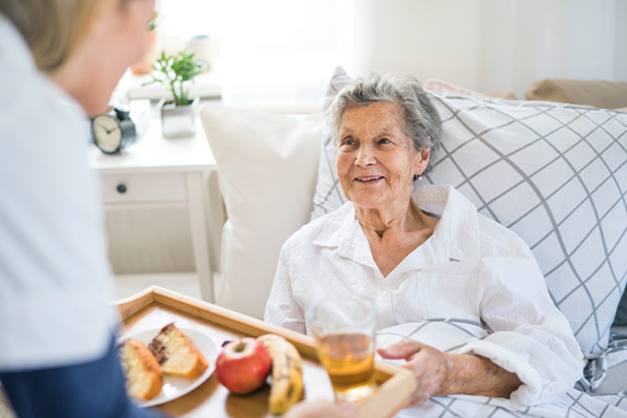 Pflegekraft bringt einer älteren Dame im Bett das Frühstück