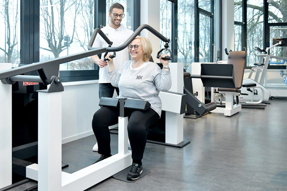 eine Frau mittleren Alters sitzt auf einem Trainingsgerät und wird von einem daneben stehenden jungen Trainer im Fitnessraum begleitet