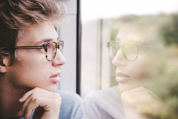 Jungendlicher mit Brille blickt aus dem Fenster und spiegelt sich in der Scheibe