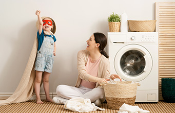 Mutter sitzt neben Waschmaschine und ein kleiner Junge steht wie Supermann verkleidet neben ihr