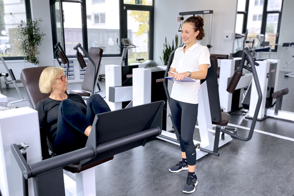 eine Frau mittleren Alters sitzt auf einem Trainingsgerät und unterhält sich mit der daneben stehenden jungen Trainerin im Fitnessraum