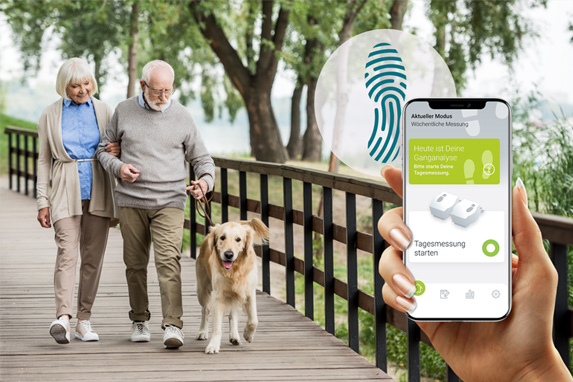  Ein Seniorenpaar spaziert mit einem Hund auf einer Holzbrücke, im Vordergrund hält eine Hand ein Smartphone mit der ParkinsonGo-App ins Bild