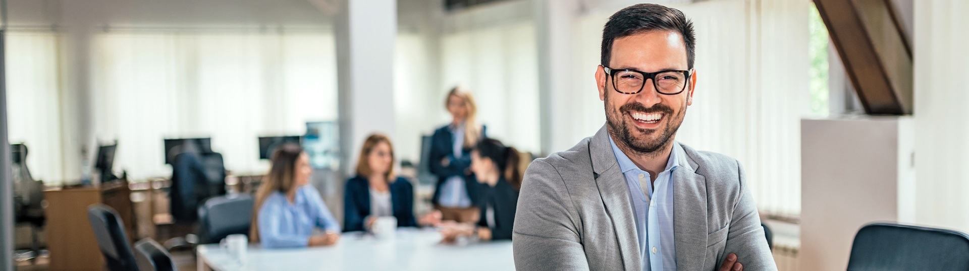 Junger Geschäftsmann mit Brille lehnt locker lächelnd an einem Besprechungstisch, im Hintergrund sitzen Kollegen am Tisch